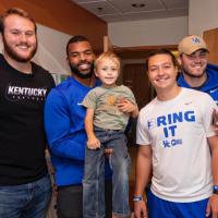 Four football players pose in a hospital room. Courtney Love, a young-looking Black man in a blue shirt, holds a young boy who is a patient at Kentucky Children’s Hospital.