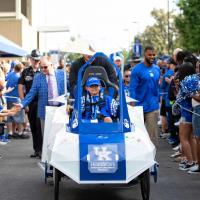 Maximo Shemwell, strapped into the cart, leads the football team down the Cat Walk between rows of cheering fans. Luke Fortner is pushing the cart. Coach Stoops and Courtney Love are visible to either side of the cart.