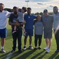 Six people pose for a photo on the football practice field. One man is holding Kase Chaney, an active kid who is holding a football. Coach Stoops, Courtney, and Luke are all in the photo, along with Kase’s parents.