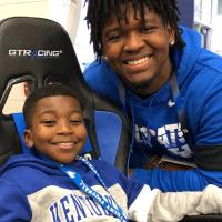 DJ Johnson, a young Black man in a Kentucky sweatshirt, poses for a close-up photo in the cart. A Black man on the UK football staff poses with him. Both are smiling.