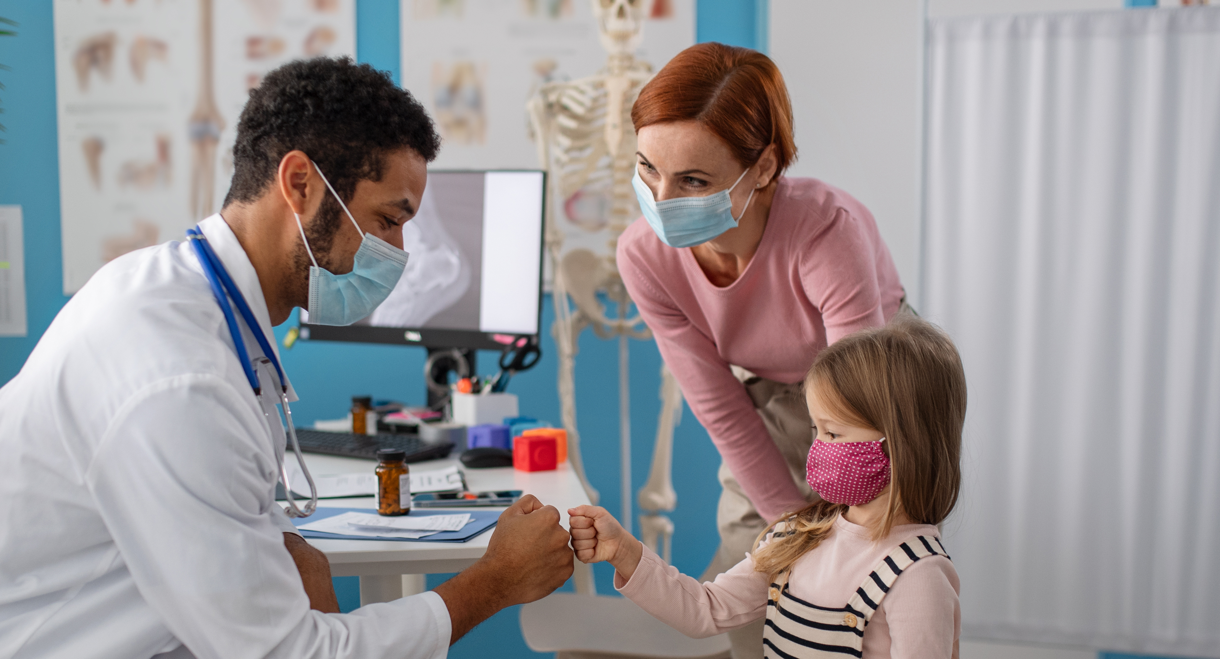 Little girl fist bumps with male doctor while her mother looks on. All three are wearing masks.