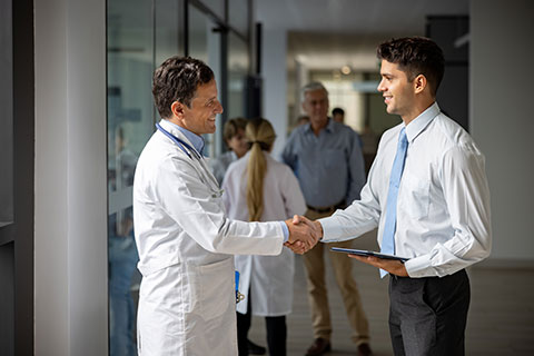 Doctor in a white coat smiles while shaking the hand of a young man in business attire