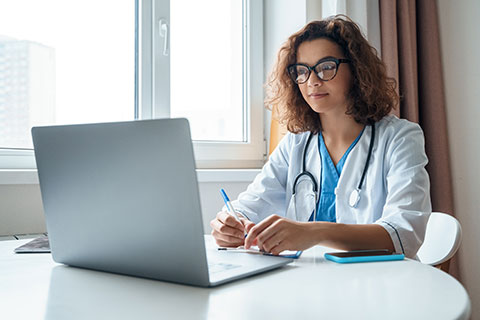 Female doctor wearing a white coat and stethoscope sitting at her laptop computer and taking notes with a pen and paper.