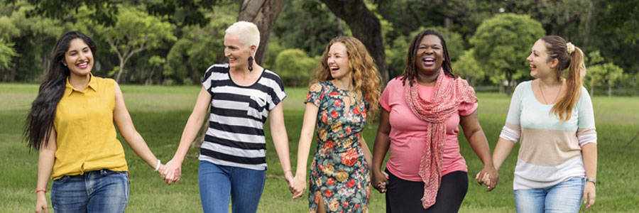 Five women of varying ages and ethnicities walk hand-in-hand across a lawn.