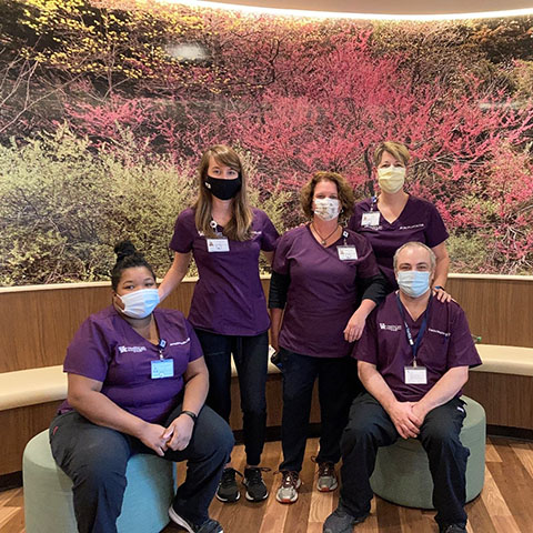 Five people wearing surgical masks, purple scrub tops and black pants in front of a mural depicting trees in springtime.