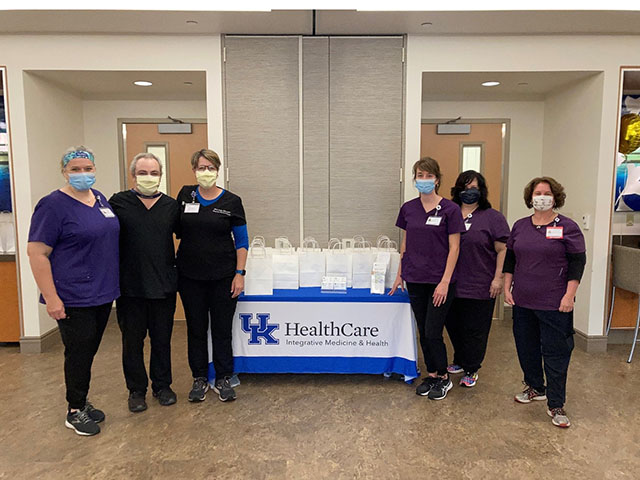 Six people in scrubs and surgical masks standing next to a table with white bags arranged on top. UK Integrative Medicine and Health is printed across the front of the tablecloth.