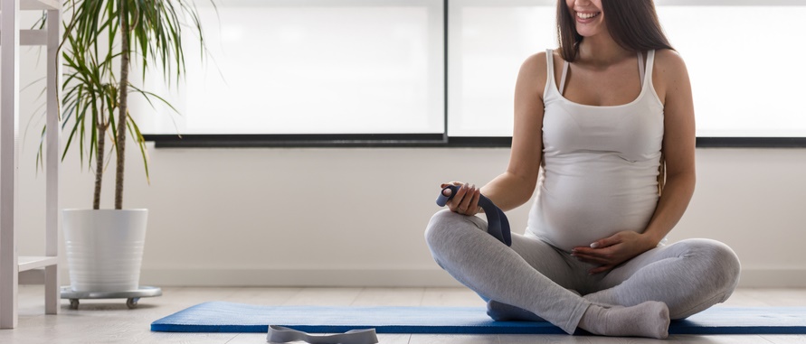 A pregnant woman sits cross-legged on a yoga mat about to stretch.