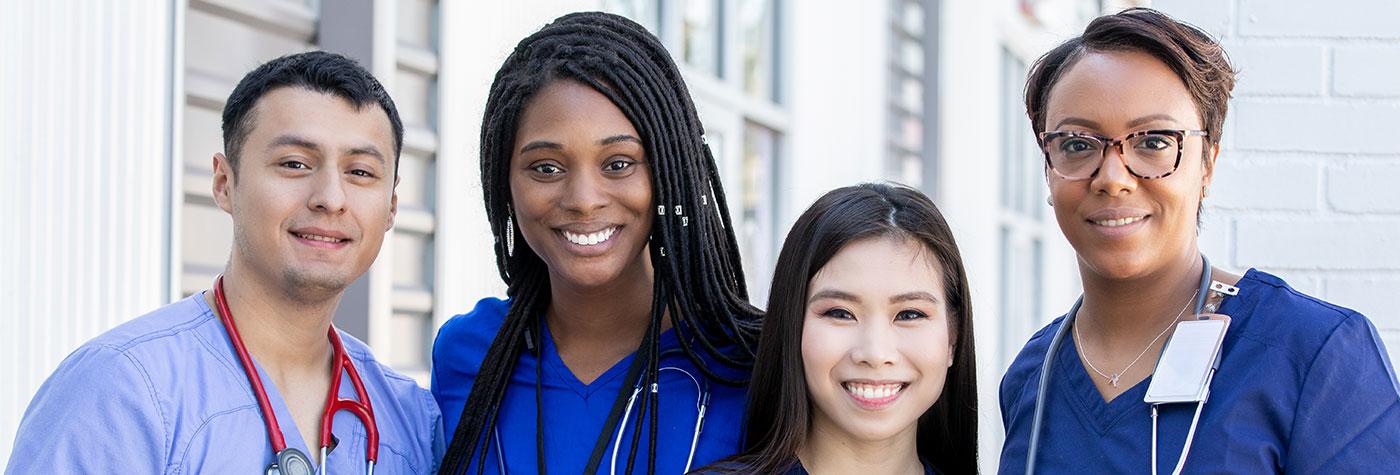 Four diverse young adults wearing scrubs with stethoscopes around their necks stand side by side, smiling and looking directly into the camera.