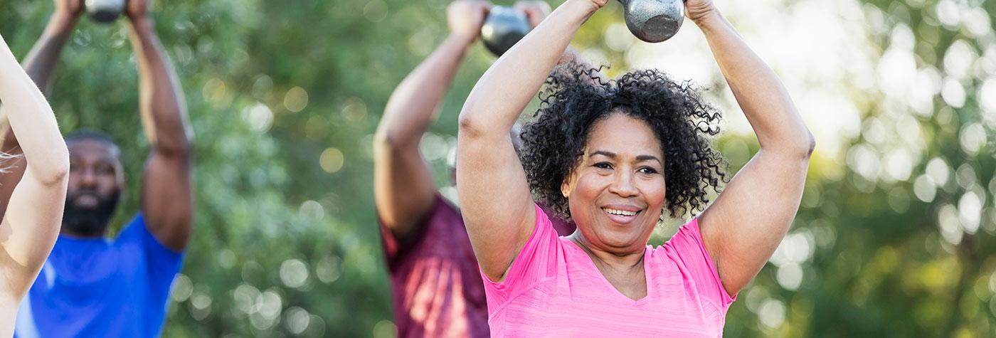 Smiling African-American woman lifts handweights as part of a group fitness class taking place outdoors.