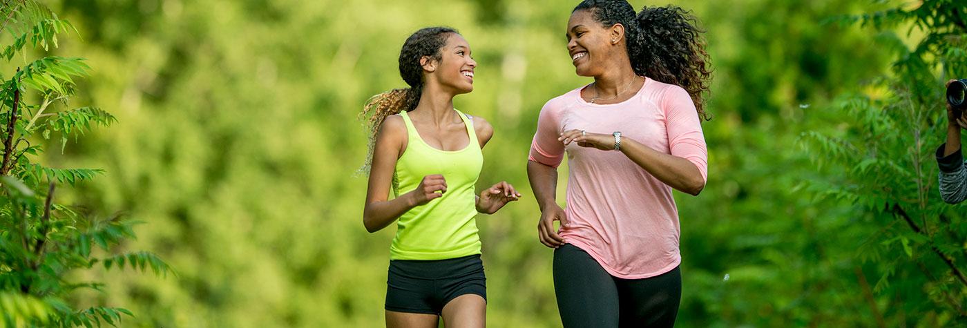 A mother and daughter are wearing athletic clothing and running through a park with trees on a sunny day. They are smiling and looking at each other.