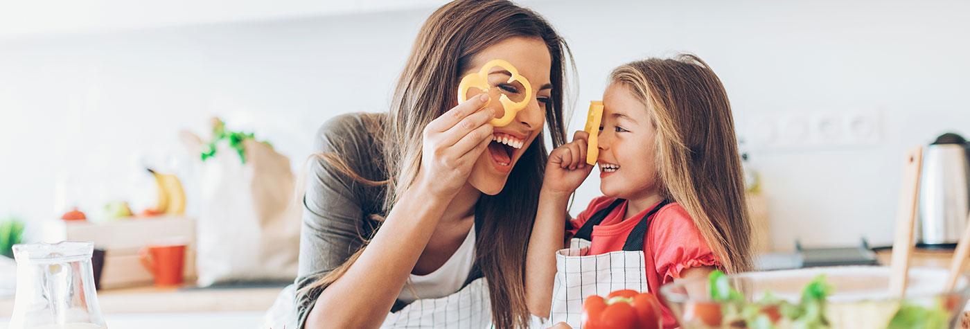 A mother and her young daughter laugh together as look through bell pepper rings while cutting vegetables for a salad.