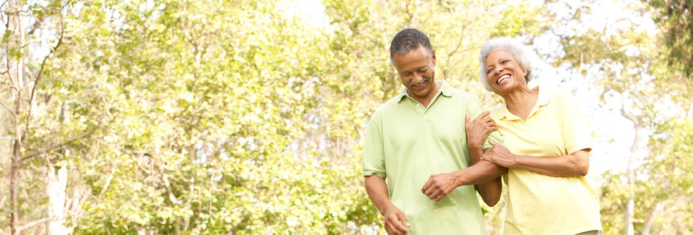 An older African-American couple smiles as they walk arm in arm through a wooded park. 