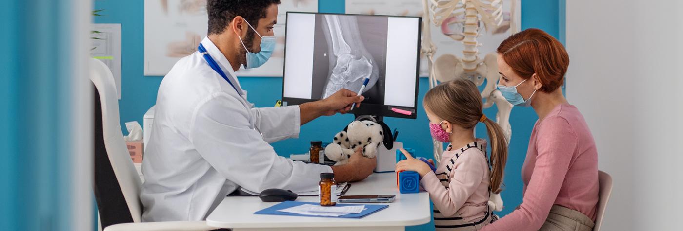 A little girl sits on her mother's lap at a doctor's desk while the doctor holds a stuffed toy and uses a pen to point to an X-ray image on his computer monitor.