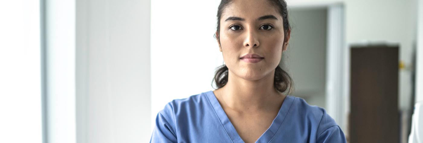 A female provider dressed in blue scrubs looks into the camera with a pleasant yet serious expression on her face. 