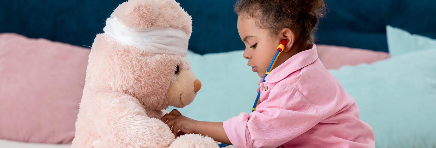 A little girl wearing a pink shirt uses a toy stethoscope to "listen" to a large teddy bear's heartbeat.