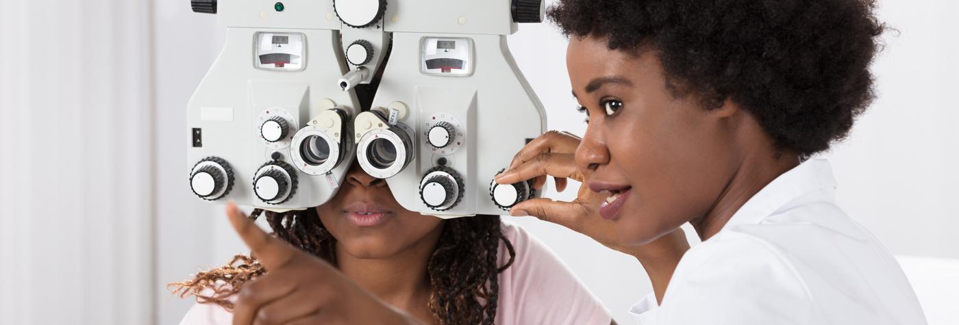 An African American female patient looks into a manual refractor while her provider, an African American woman wearing a white coat, adjusts the machine and speaks with her.