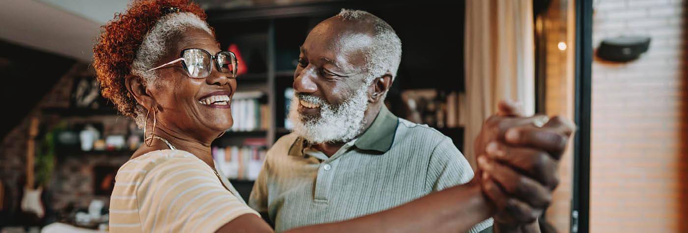 A smiling, older Black woman and a Black man with a white beard dance together.