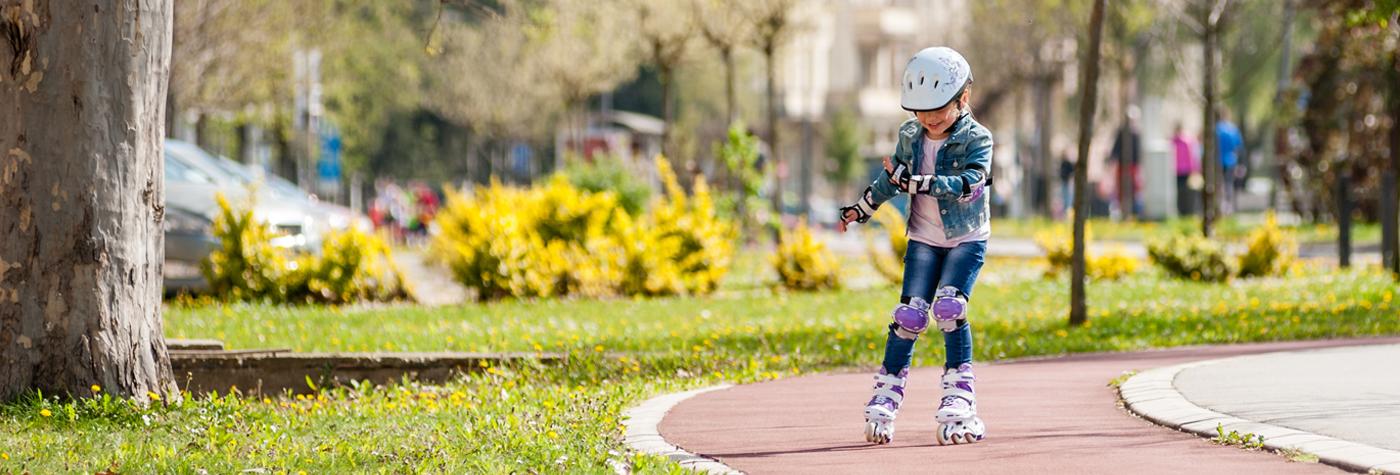 A little girl wearing kneepads, elbow pads and a helmet rollerblades in a park.