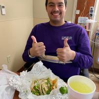 Adiel Nájera smiles at the camera and gives two thumbs up while seated at a table with a takeout order of tacos and a soft drink in front of him.