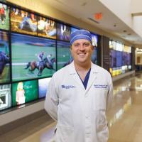 Dr. David Dornbos, a neurosurgeron at UK HealthCare, stands while smiling in front of a screen displaying race horses.