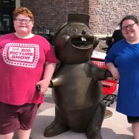 A photo of Abby and her brother, Drew, posing together with a metal statue of the Bucee’s mascot. Drew is a young white boy with curly red hair. He is wearing a short-sleeve red t-shirt that reads “The Big Picture Show” and a pair of red shorts.
