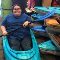 A photo from inside a sports equipment store showing Abby smiling and sitting in a kayak while pretending to row with the paddle she is holding.