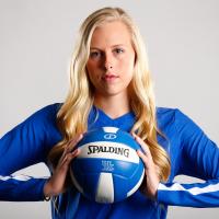 A portrait of Alli standing against a white backdrop, holding a volleyball between her palms. She looks determined and strong.