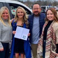 Allie smiles and poses with her family after graduation. Her mother is a white woman with blonde hair, wearing a high-neck gray ribbed sweater and blue jeans. Her father is an older white man with short dark hair, wearing a blue puffer vest, light-blue long sleeve shirt, khakis, and glasses. Her sister is a young brunette woman wearing a patterned shawl over a long sleeve black shirt and blue jeans.