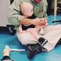 Dr. Talwalkar, an older man with salt-and-pepper scruff wearing dark-framed glasses, a green button-up, and khakis, holds Arlo in his arms as he prepares to fit him for a prosthetic.