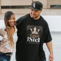 A candid photo of Brandi Jones and her partner Cory Jones walking together. She is a Black woman with long black and blonde ombre braids. She is wearing a white button-up with an ornate pattern, a pair of blue jeans, and a pair of round earrings that say “1SicB” in the middle. He is a Black man with a black fitted cap on. He is also wearing a black short-sleeve t-shirt that reads “Certified 1SicB.”
