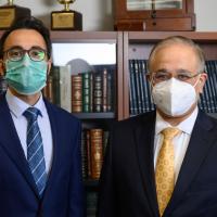 Dr. Farhan Mirza (left), a younger man with dark hair and a beard with glasses, and Dr. Siddharth Kapoor (right), a middle aged man with glasses, both dressed in suit jackets and ties with face masks on standing side by side in front of a bookshelf.