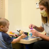 A candid photo of Child Life Specialist Sydney Houzenga and a patient playing with bubbles while sitting at a table. Sydney is a young white woman with mid-length brunette hair. She is wearing a short-sleeve gray t-shirt and a yellow medical face mask.