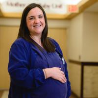 Kelsey stands smiling in the hallway at UK HealthCare. She is wearing her navy scrubs with her hand placed on her belly, showing her baby bump.