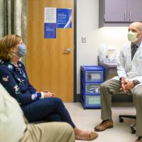 E and her husband Cary, an older white man with white hair, sit in a hospital room with Dr. Fred Ueland, a middle-aged white man with short-cropped grey hair. Dr. Ueland is wearing a white lab coat. All three are wearing masks.