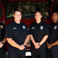 Four adult men in EMS and Fire Rescue uniforms standing with their hands clasped in front of a fire engine.