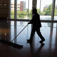 A person wearing a face mask and protective glasses mopping a tile floor. They are silhouetted against a large wall of windows.