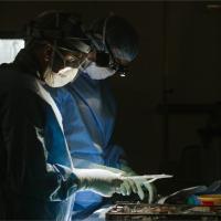 Two surgeons gather their tools for surgery. Both are wearing scrubs, masks, goggles, and gloves. The photo is dark, with dramatic lighting on their tools.