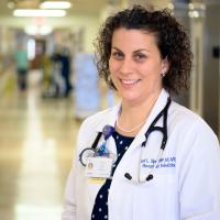 Geri stands in the hallway of the hospital. She is wearing a white coat with a stethoscope around her neck, a pearl necklace, and blue polka dot blouse.