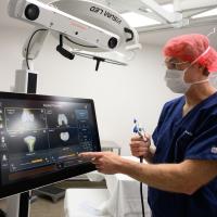 A photo of Dr. Jeffrey Selby pointing at X-rays on a screen in his operating room. He is an older white man with short gray hair. He is wearing navy scrubs, a white face mask, a salmon-colored hair net, and a pair of gold-rimmed glasses.