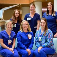 Jennifer poses and smiles alongside five of her fellow nurses, all of whom are white women wearing blue scrubs.