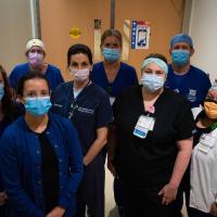 Eight UK HealthCare team members are gathered together in a hospital hallway. All are wearing masks, as well as blue, black or white scrubs.