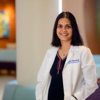 A photo of Dr. Asha Shenoi smiling for the camera with her hands in her pockets. Dr. Shenoi is a South Asian woman with mid-length black hair. She is wearing a white lab coat over a navy top that has a red ribbon around the collar.