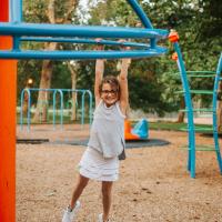 Gabriella Buffano, approximately 10 years old, holds onto a piece of playground equipment, swinging by her arms and grinning. She is wearing a white skirt, a gray tank top, and glasses.