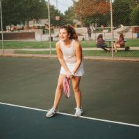 An action shot of Isabella, 12 years old, on the tennis court. She is wearing a white skirt and a gray tank top, and her curly brown hair is pulled back inn a ponytail.