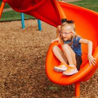 Olivia Buffano, six years old, comes down a slide. She has blonde hair with a black bow in it, and is wearing a blue Kentucky shirt, orange sneakers, a black skirt, and pink glasses.
