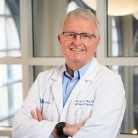 Dr. David Booth, an older white man with grey hair, stands smiling with his arms crossed over his chest. He is wearing a doctor’s coat, blue button-down, and spectacles.
