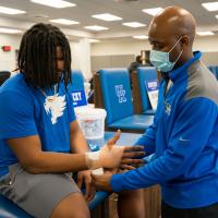 Kenneth, wearing a gold chain, blue shirt and grey shorts, sits on a UK chair as Gabe wraps his wrist with tape. Gabe is wearing a facemask over his mouth and nose.