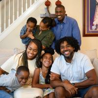The entire Love family smiles and poses for the camera in their home. Kristen’s grandfather is an older African-American man with dark short hair and a mustache, and is wearing a long-sleeve blue button-up. Kristen’s grandmother is an older African-American woman with medium-length dark hair, and she is wearing an army green top with ruched sleeves.
