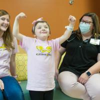 Leorah has a smile on her face and her arms in the air as she strikes a pose, while her mother Nicole and one of her nurses look on as they sit on a couch, smiling and laughing. Nicole is a white adult woman with light-brown hair and is wearing a pink “Leorah Strong” shirt. The nurse is a white adult woman with brown hair and is wearing a medical mask, a black half-button shirt, and khakis.