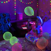 Lenorah plays with a balloon while sitting on her hospital bed, which is being illuminated by a blacklight. The room is filled with balloons and streamers, all of which are glowing.